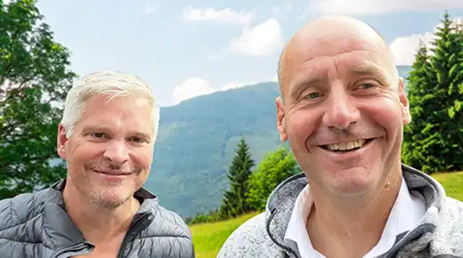 Die beiden Board-Mitglieder des Johner Instituts lächeln in einem Selfie in die Kamera. Im Hintergrund ist eine Berglandschaft zu sehen.